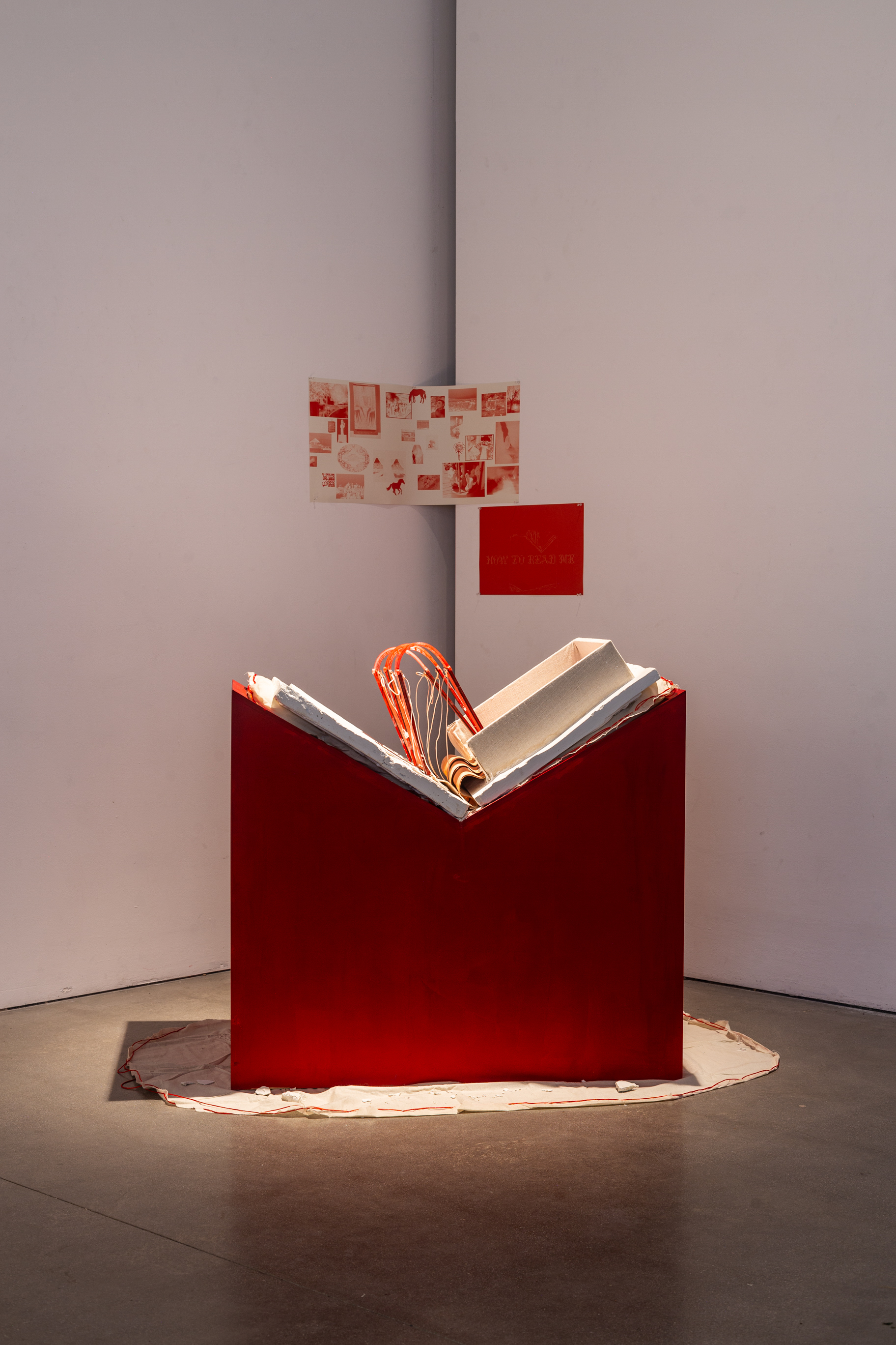 A red geometric sculpture with white and gray slabs forming a book protruding from the top, displayed against a plain white wall.